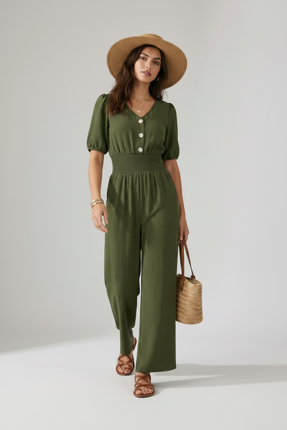A woman models an olive green short-sleeve jumpsuit with puff sleeves, a buttoned V-neck, and wide legs. She is wearing a wide-brimmed tan hat, brown strappy sandals, and holding a woven straw handbag against a neutral studio background.