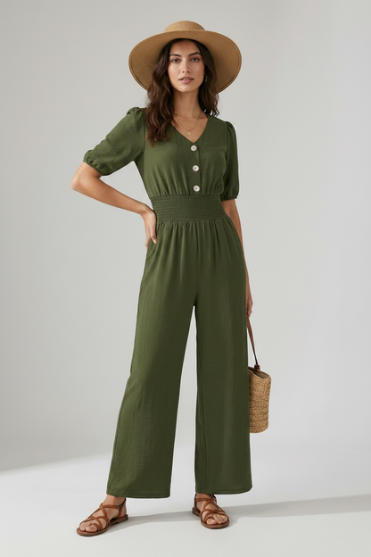 A woman models an olive green short-sleeve jumpsuit with puff sleeves, a buttoned V-neck, and wide legs. She is wearing a wide-brimmed tan hat, brown strappy sandals, and holding a woven straw handbag against a neutral studio background.