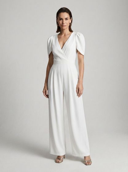 A woman modeling a white wide-leg jumpsuit with short puff sleeves, a wrap-style V-neck bodice, and a cinched waist, photographed against a light gray studio background.