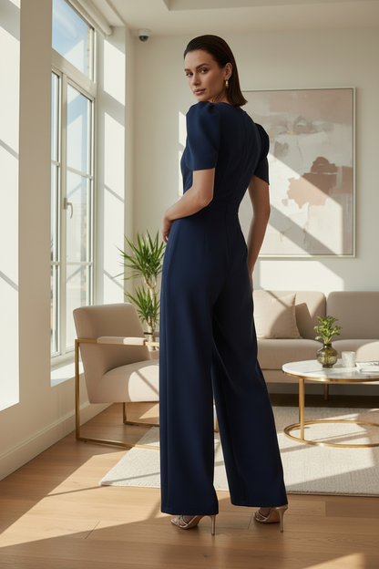 A woman stands in a bright, modern living room wearing a navy blue short-sleeve wide-leg jumpsuit, photographed from behind to show the back of the garment.