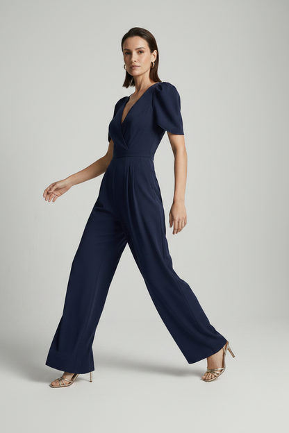 A woman modeling a navy blue jumpsuit with puff sleeves, a wrap-style V-neckline, and wide legs, photographed against a plain light gray studio background.