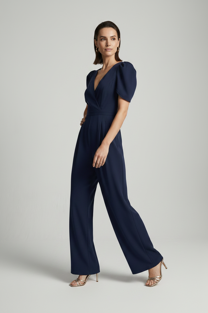 A woman modeling a navy blue jumpsuit with puff sleeves, a wrap-style V-neckline, and wide legs, photographed against a plain light gray studio background.