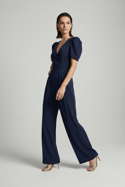 A woman modeling a navy blue jumpsuit with puff sleeves, a wrap-style V-neckline, and wide legs, photographed against a plain light gray studio background.
