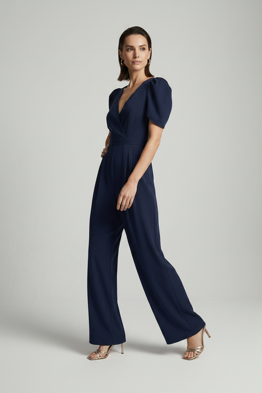 A woman modeling a navy blue jumpsuit with puff sleeves, a wrap-style V-neckline, and wide legs, photographed against a plain light gray studio background.