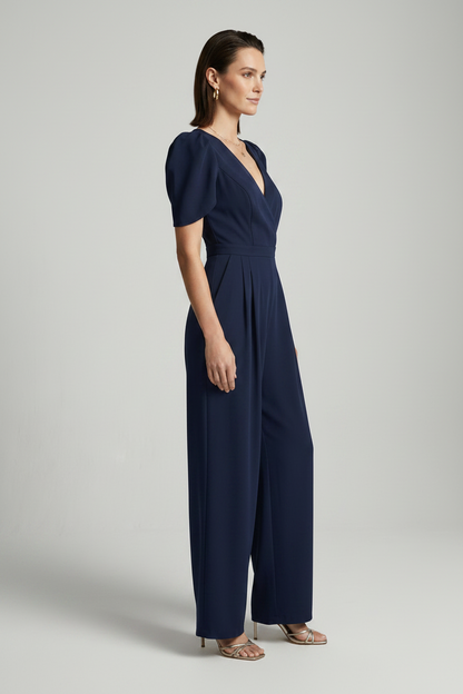 A woman modeling a navy blue jumpsuit with puff sleeves, a wrap-style V-neckline, and wide legs, photographed against a plain light gray studio background.