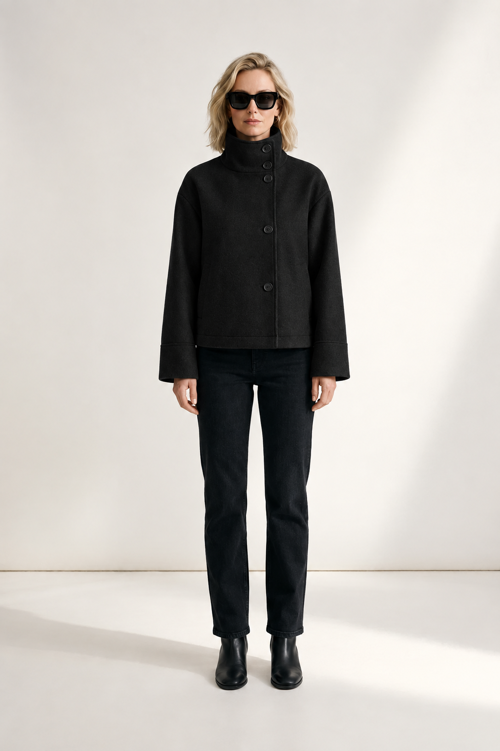 A woman modeling a gray cropped button-front jacket with a high mock neck, paired with black pants and black boots, standing against a clean off-white studio background.