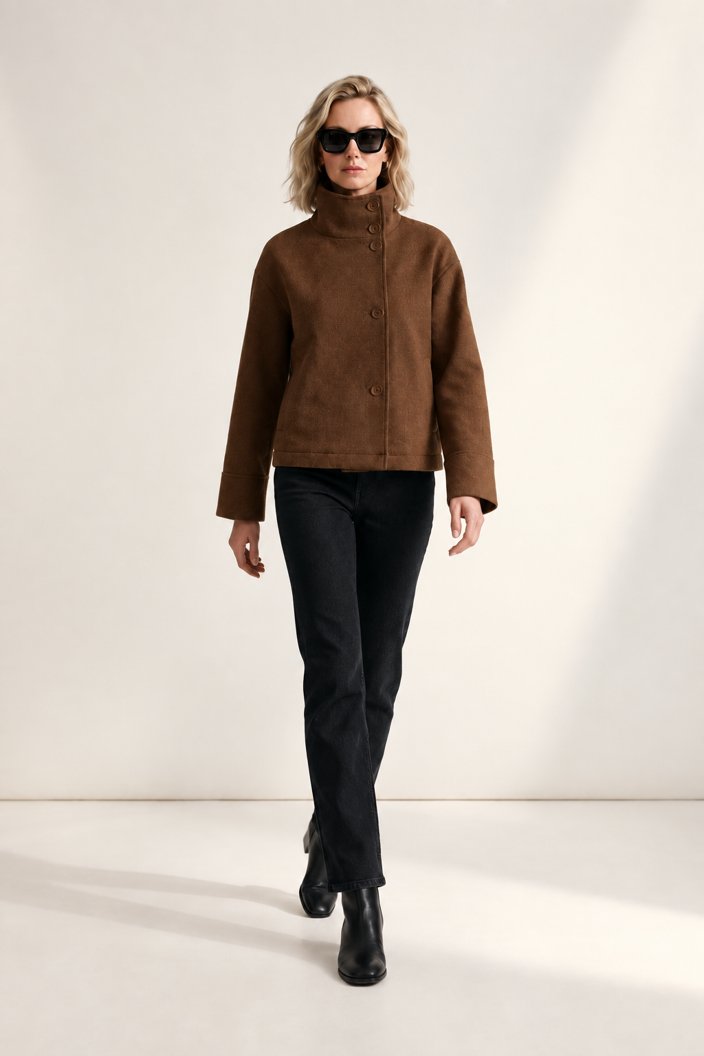 A model stands against a minimalist off-white studio background wearing a brown cropped jacket with a high mock neck and front buttons, black pants, black ankle boots, and black sunglasses.