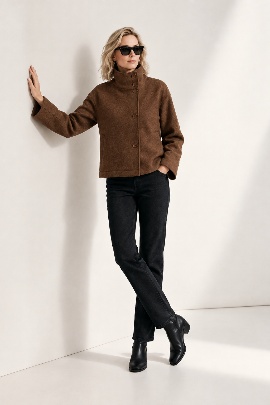 A model stands against a minimalist off-white studio background wearing a brown cropped jacket with a high mock neck and front buttons, black pants, black ankle boots, and black sunglasses.