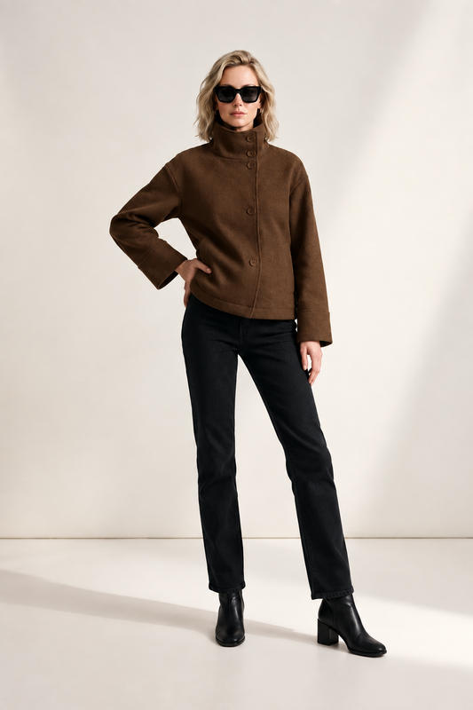 A model stands against a minimalist off-white studio background wearing a brown cropped jacket with a high mock neck and front buttons, black pants, black ankle boots, and black sunglasses.