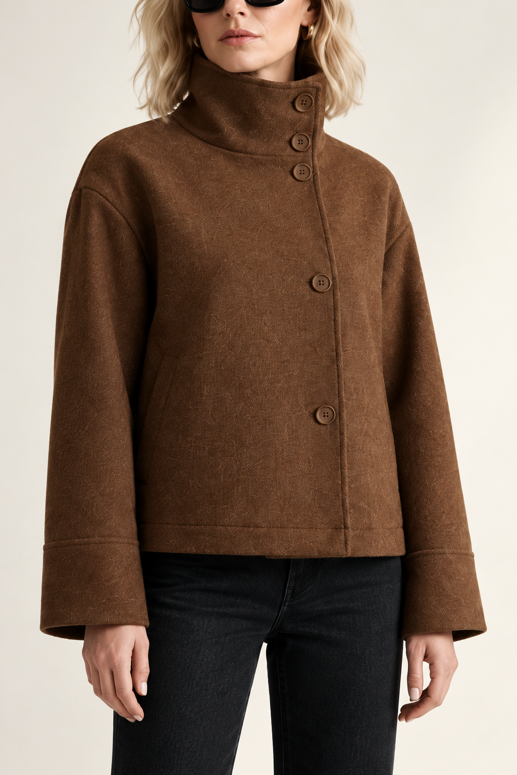 A model stands against a minimalist off-white studio background wearing a brown cropped jacket with a high mock neck and front buttons, black pants, black ankle boots, and black sunglasses.