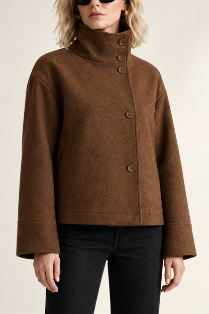 A model stands against a minimalist off-white studio background wearing a brown cropped jacket with a high mock neck and front buttons, black pants, black ankle boots, and black sunglasses.