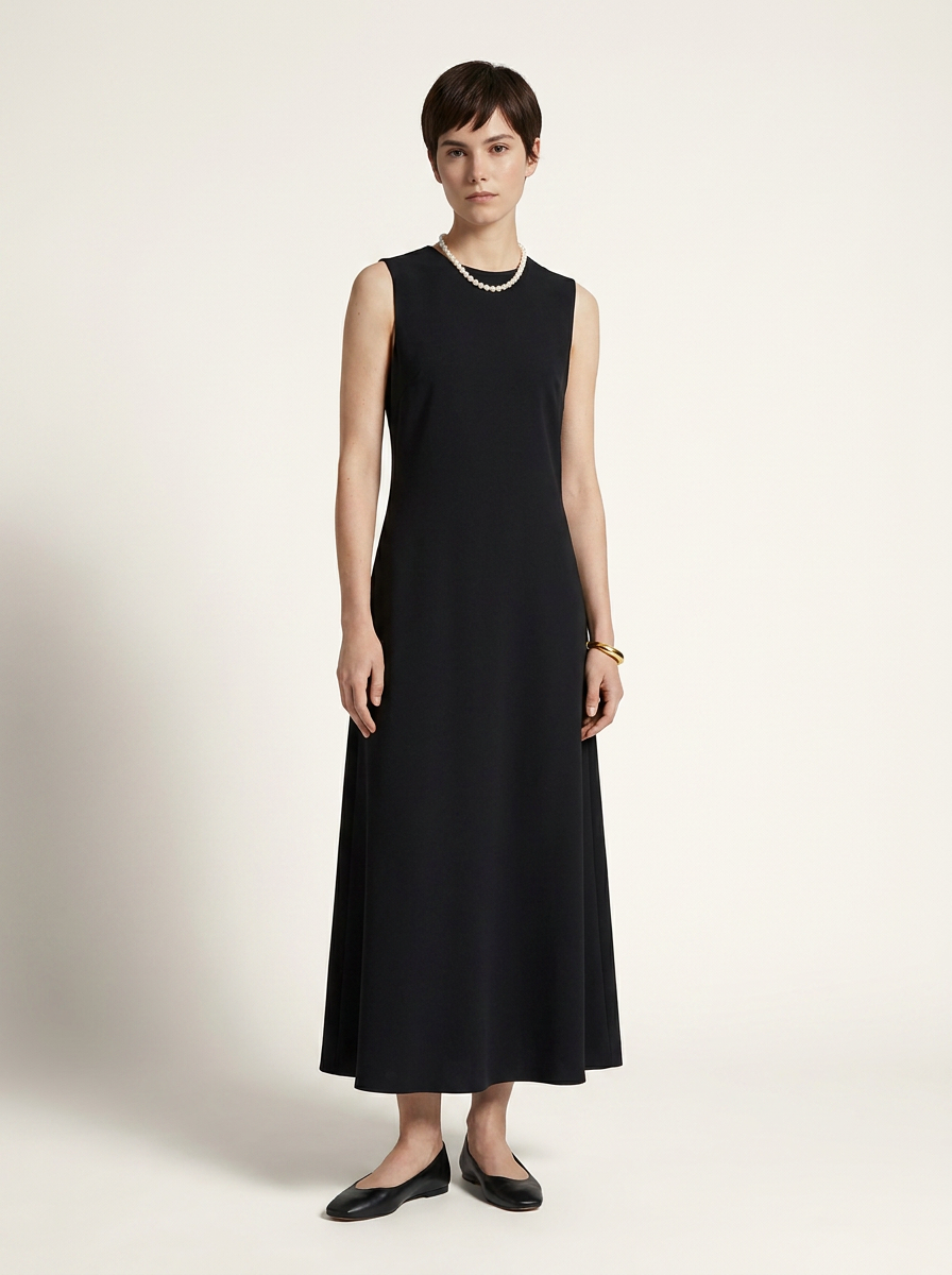A woman modeling a sleeveless black midi dress with a crew neckline, paired with black flats and a gold necklace, standing against a plain off-white studio background.
