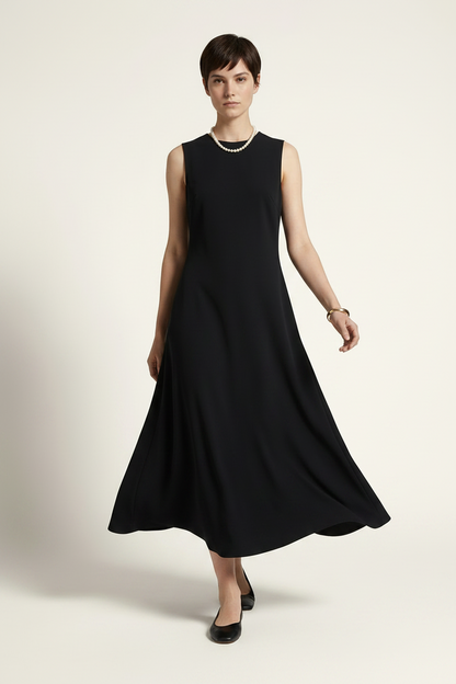 A woman modeling a sleeveless black midi dress with a flared skirt, styled with a pearl necklace and black flats against a warm off-white studio background.