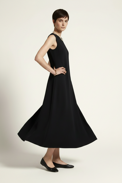 A woman modeling a sleeveless black midi dress with a flared skirt, styled with a pearl necklace and black flats against a warm off-white studio background.
