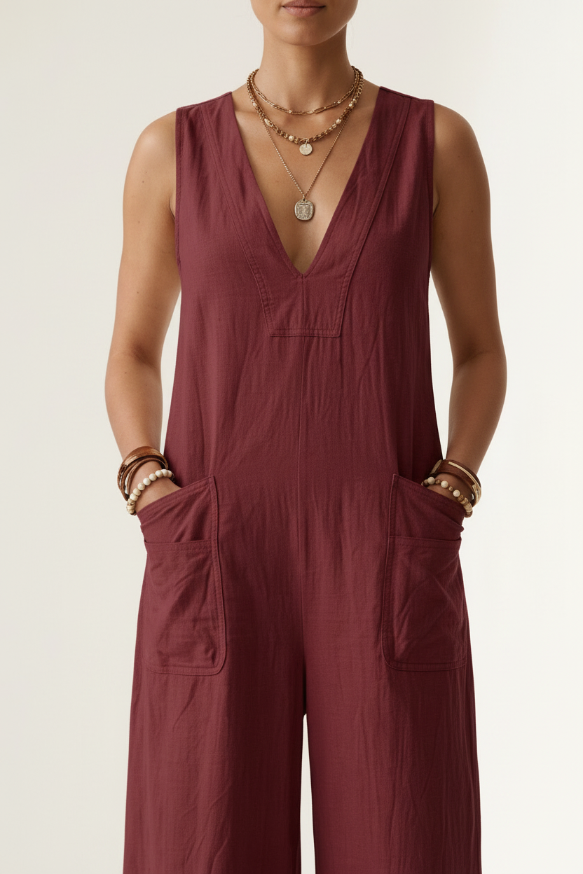 A woman stands against a plain light beige studio background wearing a sleeveless wide-leg jumpsuit in deep burgundy. She is holding a woven straw tote bag and wearing gold jewelry and brown sandals.