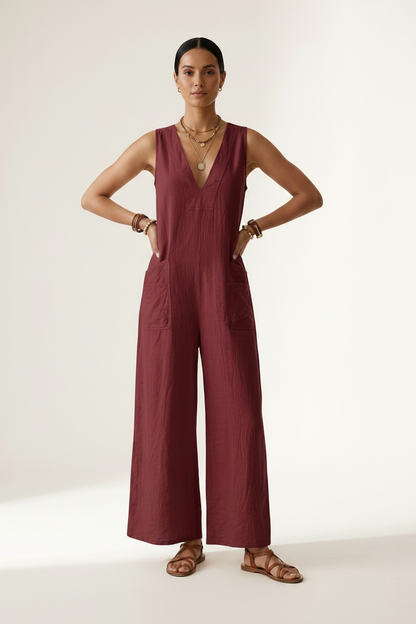 A woman stands against a plain light beige studio background wearing a sleeveless wide-leg jumpsuit in deep burgundy. She is holding a woven straw tote bag and wearing gold jewelry and brown sandals.