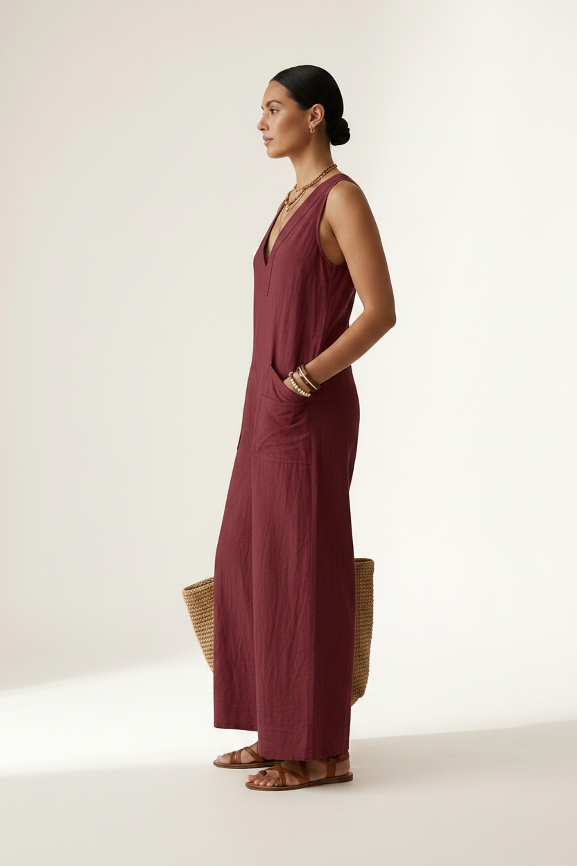 A woman stands against a plain light beige studio background wearing a sleeveless wide-leg jumpsuit in deep burgundy. She is holding a woven straw tote bag and wearing gold jewelry and brown sandals.