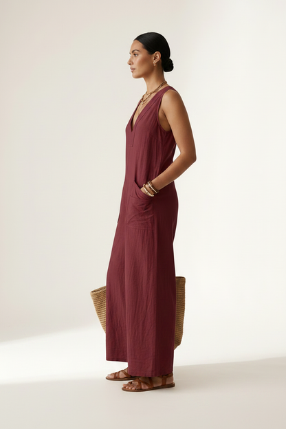 A woman stands against a plain light beige studio background wearing a sleeveless wide-leg jumpsuit in deep burgundy. She is holding a woven straw tote bag and wearing gold jewelry and brown sandals.