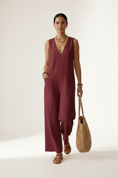 A woman stands against a plain light beige studio background wearing a sleeveless wide-leg jumpsuit in deep burgundy. She is holding a woven straw tote bag and wearing gold jewelry and brown sandals.