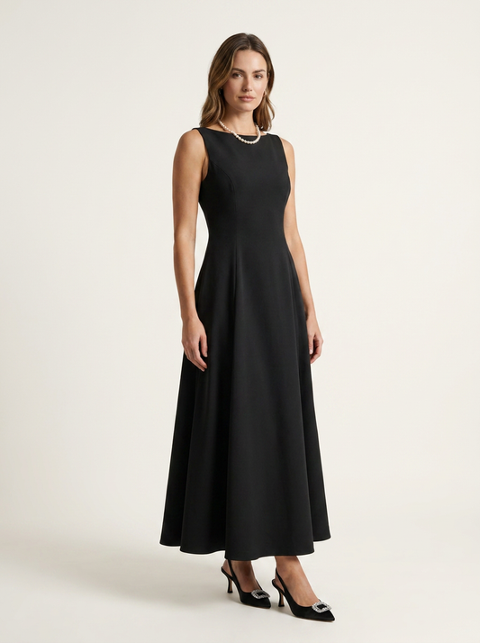 A woman modeling a sleeveless black midi dress with a fitted bodice and flared skirt, paired with black pointed-toe heels, against a plain light beige studio background.
