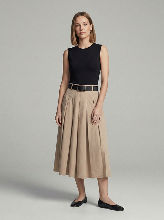 A woman stands against a light gray studio background wearing a sleeveless black top, a beige pleated midi skirt with a black belt, and black flats.