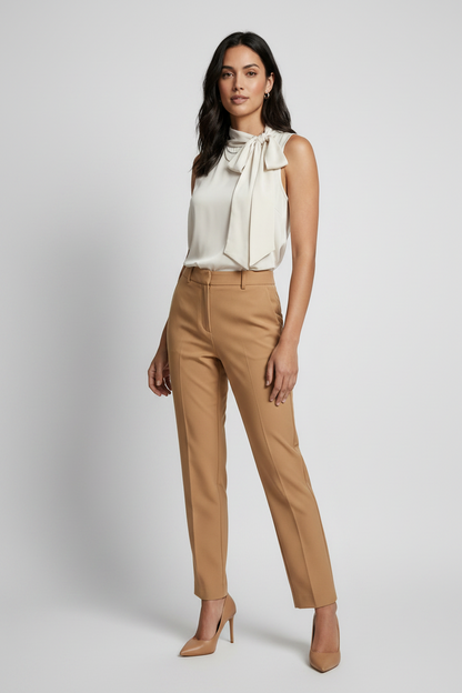 A woman modeling a sleeveless cream blouse with a large bow tie-neck detail, paired with tan trousers, against a light gray studio background.