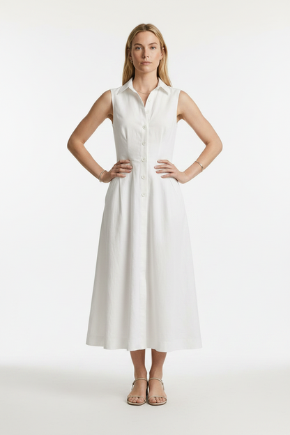 A woman stands in a studio wearing a sleeveless white midi shirt dress with a button front and side pockets, paired with beige strappy sandals against a light gray background.