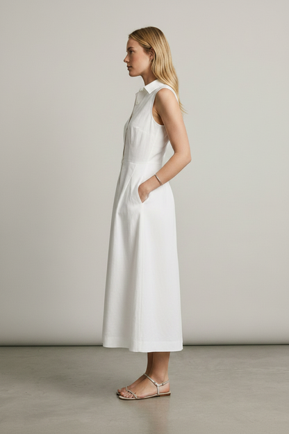 A woman stands in a studio wearing a sleeveless white midi shirt dress with a button front and side pockets, paired with beige strappy sandals against a light gray background.