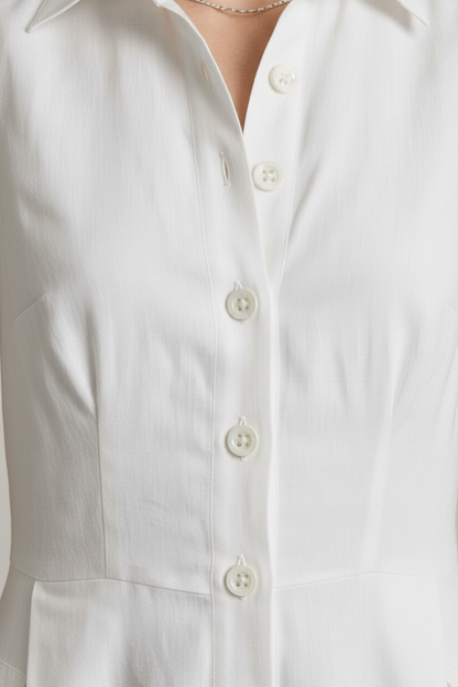 A woman stands in a studio wearing a sleeveless white midi shirt dress with a button front and side pockets, paired with beige strappy sandals against a light gray background.