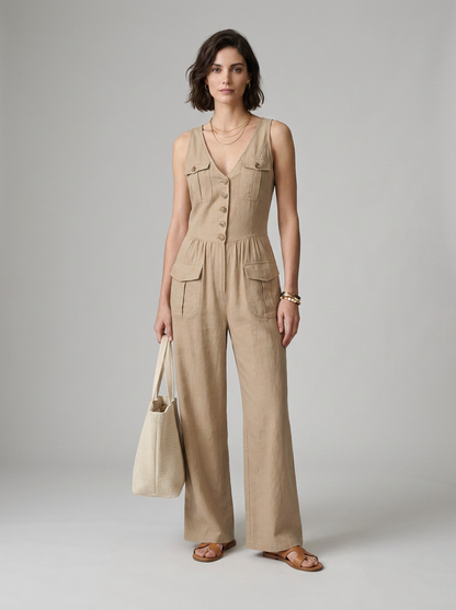 A woman models a sleeveless beige wide-leg jumpsuit with front buttons and flap pockets, holding a matching beige tote bag against a neutral gray studio background.