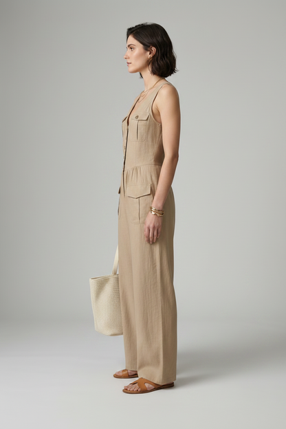 A woman models a sleeveless beige wide-leg jumpsuit with front buttons and flap pockets, holding a matching beige tote bag against a neutral gray studio background.