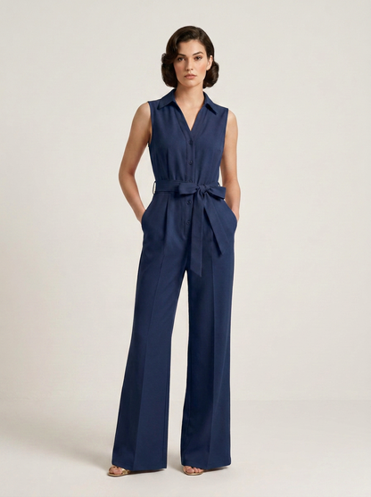 A woman modeling a sleeveless navy blue wide-leg jumpsuit with a matching waist tie, standing against a plain light beige studio background.