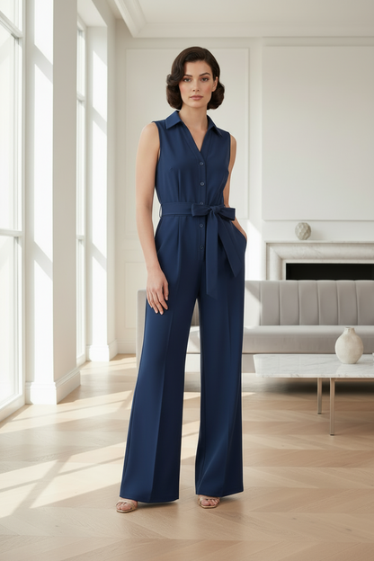 A woman stands in a bright, modern living room wearing a sleeveless navy blue wide-leg jumpsuit with a button-front bodice, waist tie, and side pockets.