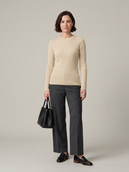 A woman stands against a plain light gray studio background wearing a cream knit sweater, dark charcoal wide-leg trousers, black loafers, and holding a black tote bag.