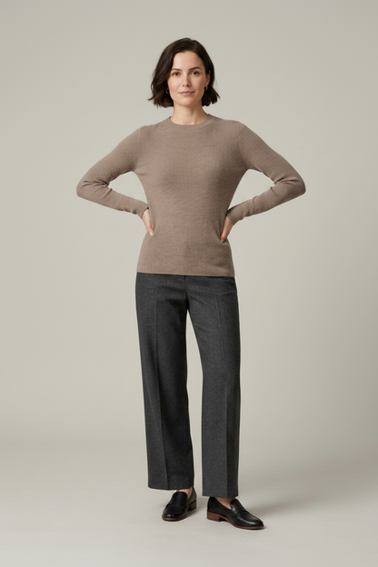A woman stands against a plain light gray studio background wearing a taupe knit sweater, dark charcoal wide-leg trousers, black loafers, and holding a black tote bag.