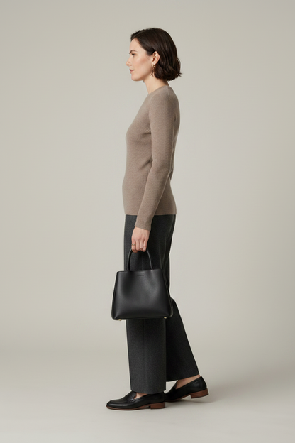 A woman stands against a plain light gray studio background wearing a taupe knit sweater, dark charcoal wide-leg trousers, black loafers, and holding a black tote bag.