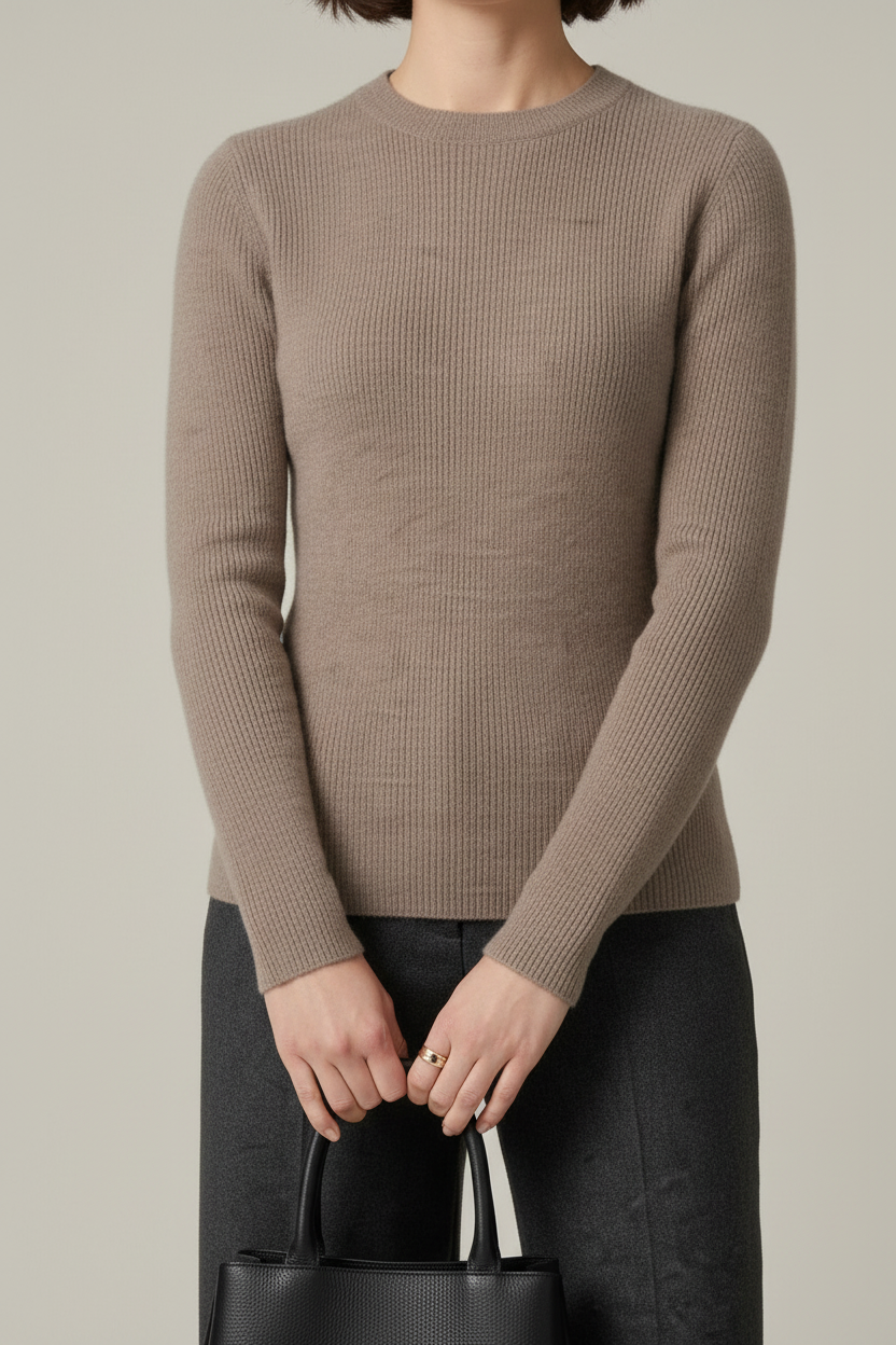 A woman stands against a plain light gray studio background wearing a taupe knit sweater, dark charcoal wide-leg trousers, black loafers, and holding a black tote bag.
