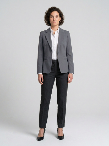 A woman modeling a tailored gray blazer with a white button-up shirt, dark charcoal trousers, and black heels against a light gray studio background.