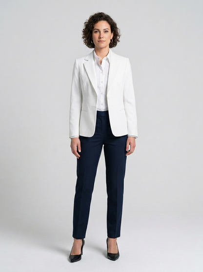 A woman stands against a light gray studio background wearing a white blazer over a white collared shirt, navy blue pants, and black pointed heels.