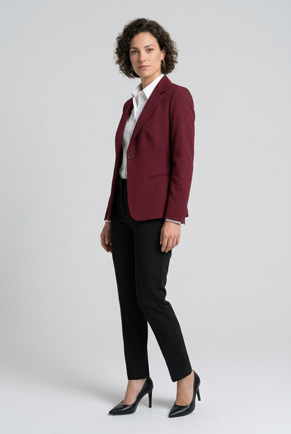 A woman stands against a light gray studio background wearing a tailored burgundy blazer over a white collared shirt, black trousers, and black heels.