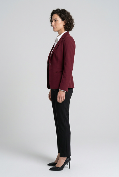 A woman stands against a light gray studio background wearing a tailored burgundy blazer over a white collared shirt, black trousers, and black heels.