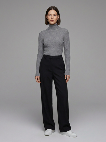 A model stands against a neutral gray studio background wearing a fitted gray ribbed turtleneck sweater, black wide-leg trousers, and white sneakers.