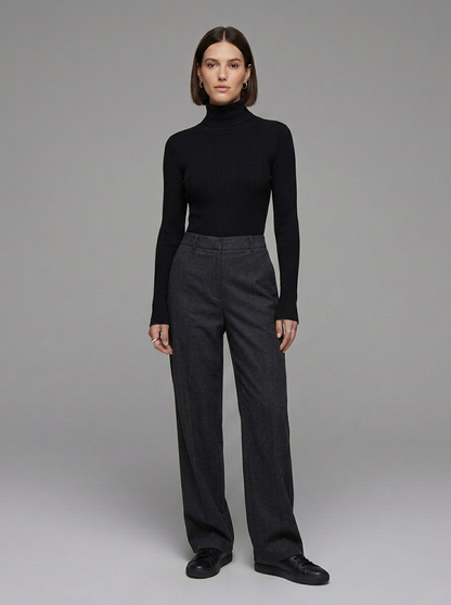 A model stands against a neutral gray studio background wearing a black ribbed turtleneck sweater, dark charcoal gray wide-leg trousers, and black leather shoes.