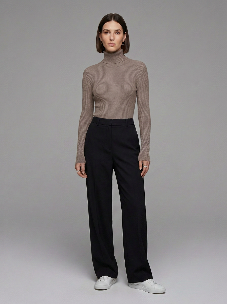 A model stands against a neutral gray studio background wearing a ribbed taupe-brown turtleneck sweater, black wide-leg trousers, and white sneakers.