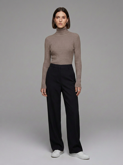 A model stands against a neutral gray studio background wearing a ribbed taupe-brown turtleneck sweater, black wide-leg trousers, and white sneakers.