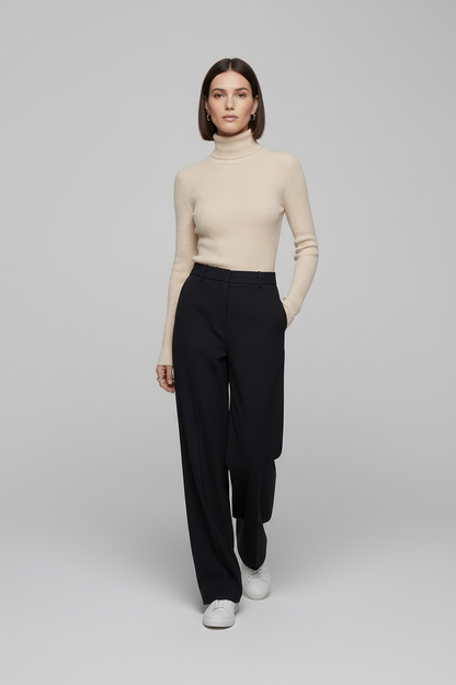 A model stands against a light gray studio background wearing a beige ribbed turtleneck sweater, black wide-leg trousers, and white sneakers.