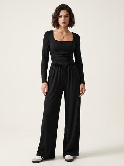 A woman modeling a black long-sleeve jumpsuit with a square neckline, gathered waist, and wide legs, standing against a plain light beige studio background.
