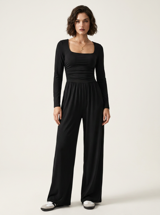 A woman modeling a black long-sleeve jumpsuit with a square neckline, gathered waist, and wide legs, standing against a plain light beige studio background.