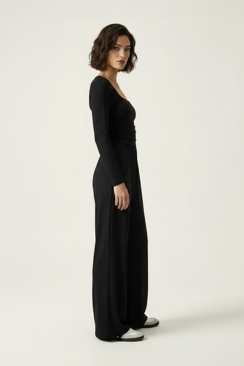 A woman modeling a black long-sleeve jumpsuit with a square neckline, ruched waist, and wide legs, paired with white slip-on shoes, against a plain light beige studio background.