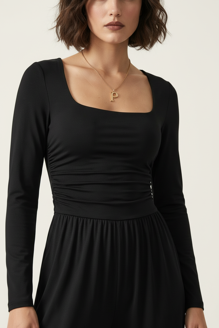 A woman wearing a black long-sleeve dress with a square neckline and gathered waist, paired with a delicate gold necklace with a letter P pendant, against a light off-white background.